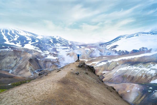 A person standing on a cliff in front of a mountain range in the Icelandic Highlands.