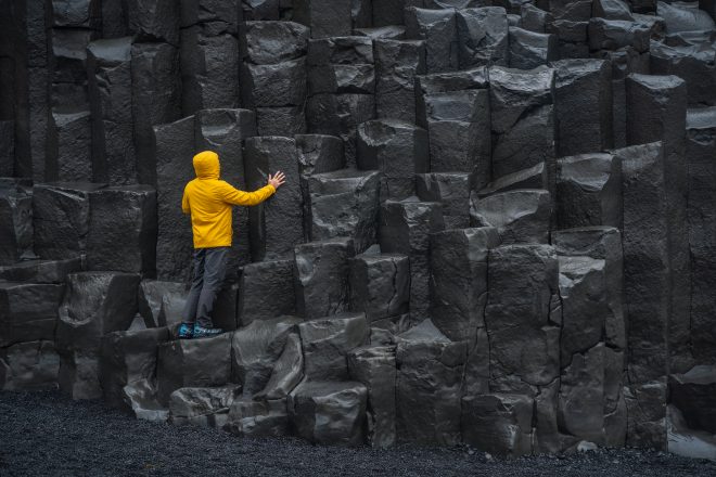 Homme adulte en veste jaune debout sur des colonnes de basalte sur la plage de sable noir de Reynisfjara en Islande