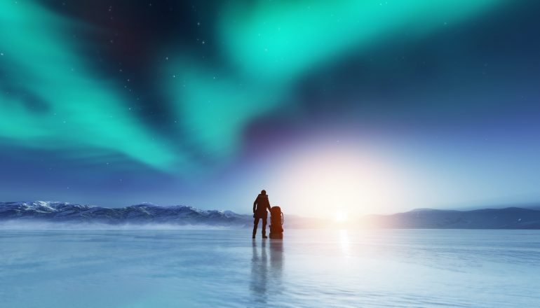 Adventurous man standing on a frozen lake with a backpack, looking at northern lights.
