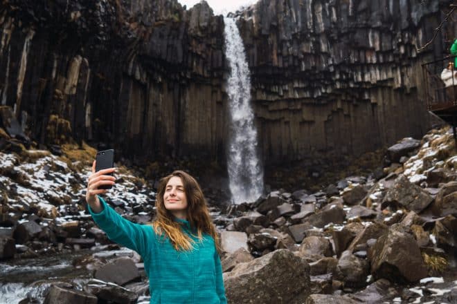 Une femme debout devant la cascade de Svartifoss en Islande, prenant un selfie sur un smartphone